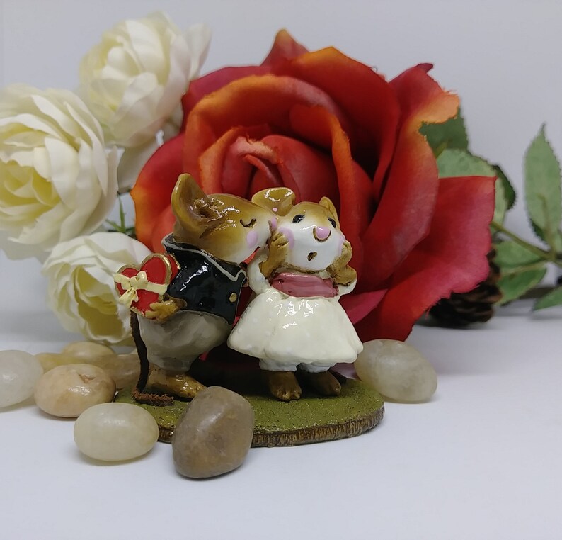May include: A figurine of two mice in formal attire, one holding a heart-shaped box. The female mouse wears a white dress and pink sash. A large red rose and white roses are in the background, with small stones in the foreground.