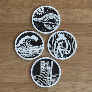 Puede incluir: Juego de cuatro posavasos redondos con diseños en blanco y negro. Cada posavasos presenta un gráfico diferente: una ola, un astronauta, un paisaje urbano y un diseño espacial. Los posavasos están hechos de un material duradero.
