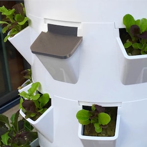 Puede incluir: Torre de jardín vertical blanca con múltiples niveles para plantar. Cada nivel tiene una maceta blanca con una pequeña planta verde que crece en ella. La torre tiene un depósito de agua gris con un pequeño pico.