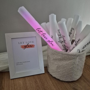 Könnte beinhalten: Ein weißer Bilderrahmen mit den Worten "LET LOVE glow" und einem rosa-orangenen Design. Ein leuchtender rosa Leuchtstab mit den Worten "let love glow" befindet sich neben einem Korb voller weißer Leuchtstäbe. Der Korb besteht aus einem strukturierten, hellgrauen Stoff.