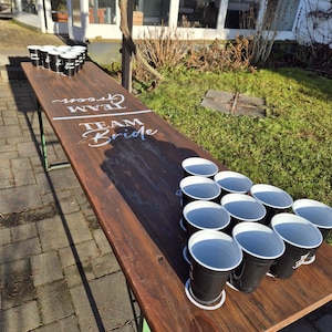 Op de afbeelding: Een houten bierpongtafel met de woorden "TEAM GROOM" en "TEAM BRIDE" in witte letters. Zwarte bekers met witte randen zijn op de tafel geplaatst. De tafel staat buiten op een zonnige dag.