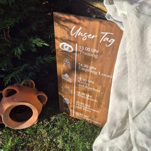 Op de afbeelding: Een houten trouwboord met witte tekst en illustraties. Het bord toont de woorden "Unser Tag" en een schema. Een terracotta pot en groen zijn op de voorgrond te zien, met een gedrapeerde stof aan de rechterkant.