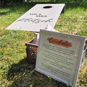 Könnte beinhalten: Ein helles Cornhole-Board mit einem kreisförmigen Loch und der Aufschrift MR & MRS Klütsch, dazu ein weiß gerahmtes Schild mit Spielanleitung und eine braune Kunststoffkiste, die mehrere Stoff-Wurfsäckchen für das Spiel enthält.