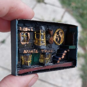 Może przedstawiać: Miniaturowa diorama wnętrza budynku, wykonana w pudełku po zapałkach. Scena przedstawia ciemną fasadę ze złotymi akcentami, małymi oknami i schodami. Na ścianie wisi okrągły obraz w złotej ramie.