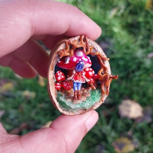 Op de afbeelding: Een miniatuur fee-figuur dat onder een rode en witte paddenstoel met witte stippen staat, omringd door kleinere rode paddenstoelen, allemaal in een uitgeholde walnootschelp.