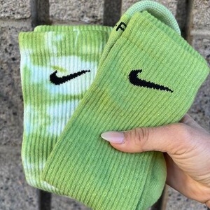 Puede incluir: Un par de calcetines verdes y blancos con un logotipo Nike swoosh. Un calcetín es tie-dye y el otro es verde sólido.