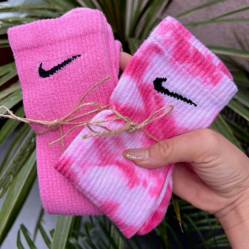 Puede incluir: Dos pares de calcetines, uno rosa y otro rosa tie-dye con un patr&oacute;n de remolino blanco. Ambos calcetines tienen un logotipo de Nike negro en forma de swoosh.