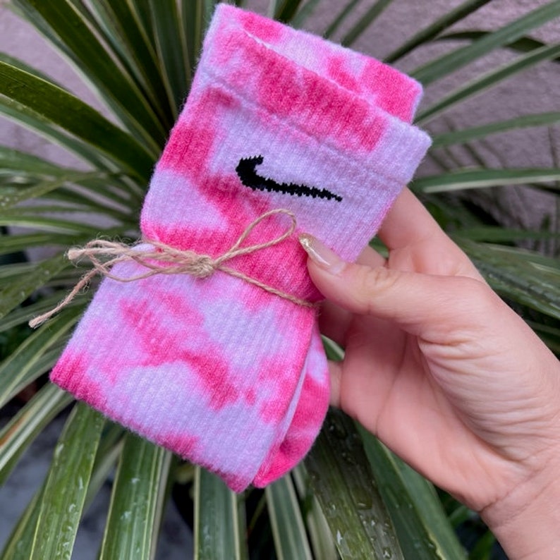 Puede incluir: Un par de calcetines de color rosa y blanco con un logotipo de Nike negro. Los calcetines est&aacute;n atados con una cuerda.