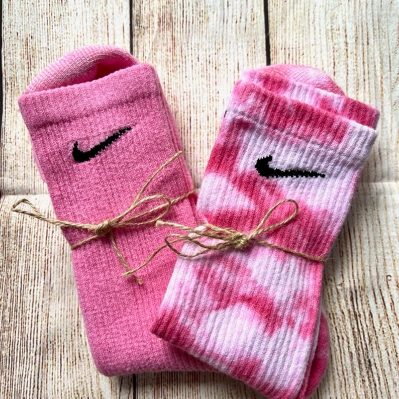 Puede incluir: Dos pares de calcetines rosas con un logotipo de Nike negro en forma de swoosh. Un par es rosa s&oacute;lido y el otro es tie-dye.