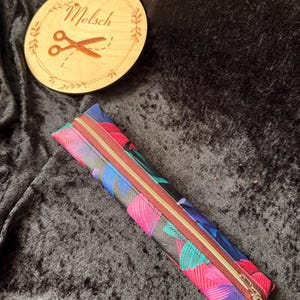 Könnte beinhalten: Ein farbenfrohes Federmäppchen mit einem Blumenmuster in Pink, Blau und Türkis, mit einem braunen Reißverschluss. Im Hintergrund befindet sich ein Holzrund mit dem Namen "Melsch" und einer Schere.