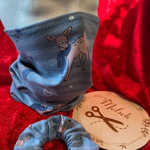 Könnte beinhalten: Eine blaue und weiße gemusterte Stoffmaske mit einem Hirsch-Design. Die Maske befindet sich auf einem Mannequin-Kopf. Ein passendes blaues und weißes gemustertes Stoffscrunchie befindet sich neben der Maske. Ein Holz-Kreis mit dem Text "Melsch" und einer Schere befindet sich im Hintergrund.