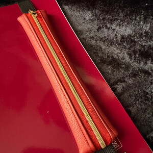 Könnte beinhalten: Ein rotes Leder-Federmäppchen mit goldenem Reißverschluss, befestigt an einem roten Notizbuch mit einem schwarzen Gummiband. Das Federmäppchen ist rechteckig und hat eine strukturierte Oberfläche.