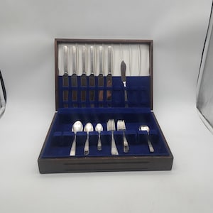 May include: A vintage silverware set in a wooden case with a blue velvet interior. The set includes knives, forks, spoons, and a butter knife. The silverware is silver-colored and arranged neatly in the case.