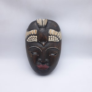Peut inclure: Un masque en bois sculpté avec une finition marron foncé. Le masque présente un visage serein avec les yeux fermés, un petit nez et des lèvres rouges. Le haut du masque comporte un élément décoratif avec des accents de couleur crème. Le masque est probablement à des fins décoratives.