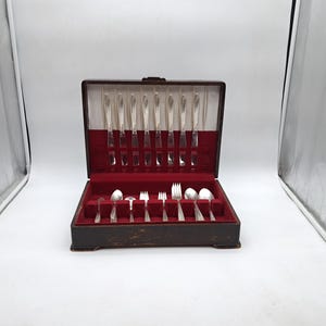 Puede incluir: Un estuche de cubiertos antiguo abierto, con interior de terciopelo rojo. El estuche contiene varios cuchillos, tenedores y cucharas plateadas. El estuche de madera es marrón oscuro, de forma rectangular y con tapa abatible.