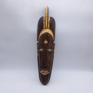 May include: A carved wooden mask with a dark brown finish. The mask features a stylized face with almond-shaped eyes, a prominent nose, and full lips. Accents of cream-colored paint highlight the eyes, mouth, and decorative elements.
