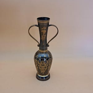 Puede incluir: Un jarrón de metal oscuro con detalles dorados y dos asas curvas. El jarrón presenta intrincados diseños florales y una base acampanada. La parte superior e inferior están adornadas con oro. El jarrón mide aproximadamente 25 cm de alto.