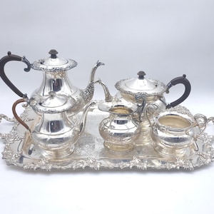 Puede incluir: Un juego de té plateado con detalles ornamentados, que incluye una tetera, una crema, un azucarero y una bandeja de servicio. La tetera tiene un asa marrón oscuro. El juego se exhibe sobre un fondo blanco.