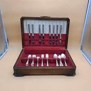 Puede incluir: Un estuche de madera para cubiertos con tapa abatible, que presenta un exterior marrón oscuro y un interior forrado de terciopelo rojo. El estuche contiene un juego de cuchillos, tenedores y cucharas de metal plateado en compartimentos.