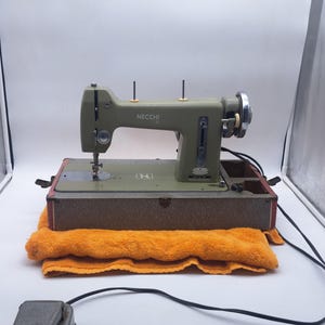 May include: An olive green Necchi sewing machine with the brand name visible. The machine sits on a brown case, atop an orange towel. A black power cord and foot pedal are in the foreground. The machine has a silver hand wheel and needle.