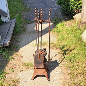 May include: A vintage wrought iron fireplace tool set. The set includes a stand with a decorative base, and tools with ornate handles. The tools are a poker, shovel, and brush. The set has a rustic, aged appearance.
