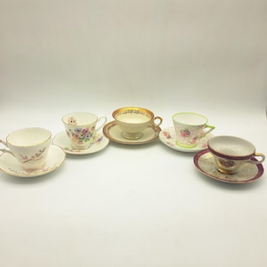 May include: Five antique teacups and saucers are arranged. The teacups showcase diverse floral and gold trim patterns. One cup has a pink floral design, another has a gold rim, and a third has a green handle. The saucers complement the teacups with matching designs.