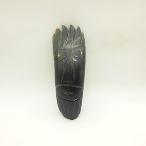 May include: A dark, carved wooden mask with a stylized face. The mask features a prominent forehead, a triangular nose, and a small mouth. The eyes are represented by small, round, orange-coloured accents. The mask is approximately 30 cm long.