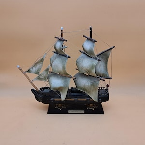 Puede incluir: Un modelo de barco con casco negro y velas de tela verde pálido desgastadas, unidas a mástiles de madera con aparejos finos. La base es negra e incluye una pequeña placa rectangular con el texto THE MOST BLESSED TRINITY impreso.