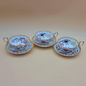May include: Three vintage teacups and saucers. The cups and saucers are white with a floral design in pink, yellow, and blue. The rims are scalloped and trimmed in gold. The handles are gold.