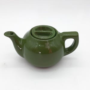 May include: A small, olive green ceramic teapot with a matching lid and a curved spout. The handle is a rectangular loop. The teapot is on a white background. This item is suitable for brewing tea.