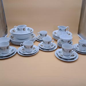 May include: A white porcelain tea set with a leaf pattern in brown and green. The set includes cups, saucers, bowls, and cream jugs. The tea set is arranged on a light brown surface.