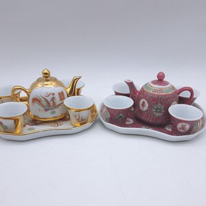 May include: Two decorative tea sets. One set features a gold teapot and cups with a rooster design. The other set has a red teapot and cups with floral patterns. Both sets include a tray.