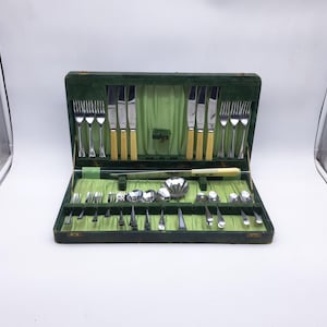 May include: An open, vintage green cutlery case with a complete set of silverware. The set includes forks, knives, spoons, and serving utensils. The handles are a light cream color, and the case has a green velvet interior.