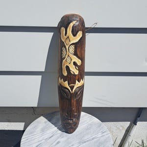 May include: A dark brown, carved wooden mask with a stylized face and abstract designs. The mask features a long, oval shape with a detailed face at the bottom and a series of carved patterns in cream-coloured wood. The mask is likely for decorative purposes.