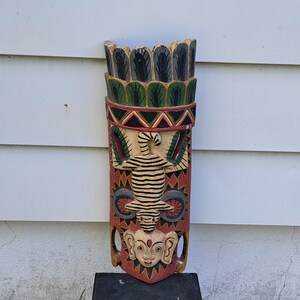 May include: A carved wooden tribal mask with a detailed design. The mask features a central figure with a striped body and a face at the bottom. The colour palette includes red, green, black, and beige. The mask is displayed on a black stand.
