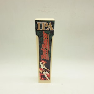 May include: A cream-coloured beer tap handle with the text "IPA" in gold letters at the top. The handle features the words "Red Racer" in red and a graphic of a woman on a bicycle. The handle is rectangular with rounded edges.
