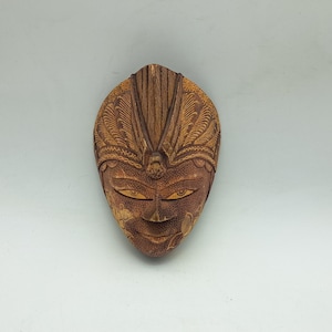 May include: A carved wooden mask with a detailed design. The mask is brown with lighter brown accents and features a stylized face with almond-shaped eyes and a small, smiling mouth. The top of the mask has a feathered design.