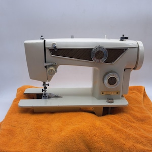 May include: An off-white sewing machine featuring a wood-grain patterned panel, two circular control dials with silver accents, and a metal needle assembly. The machine has the brand name OMEGA printed in gold lettering on the front lower right side.