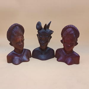 May include: Three carved wooden busts with detailed facial features and hairstyles. The busts are in varying shades of brown and red, with the central figure wearing a headdress. The background is a solid, light tan color.
