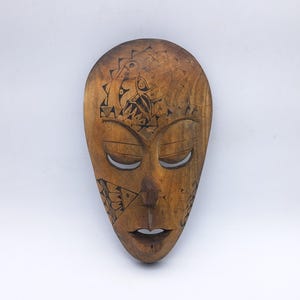 May include: A carved wooden mask with a smooth, medium-brown finish. The mask features elongated, almond-shaped eyes and a small, open mouth. Intricate black line art patterns are etched into the forehead and cheek areas of the face.