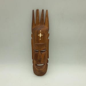 May include: A carved wooden mask with a stylized face and a crown-like top. The mask is a warm brown colour with inlaid white accents. The face has detailed features, including a smiling mouth and carved lines.