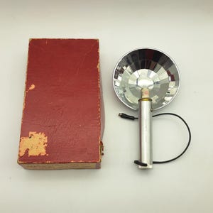 May include: Vintage camera flash unit with a silver reflector and a silver body. The flash is next to a red cardboard box with some wear and tear. A black cord extends from the flash unit.