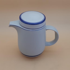 May include: A light grey ceramic teapot with a white lid and blue trim. The teapot has a curved handle and a spout. The design includes a blue stripe around the base and the lid. The teapot is approximately 18 cm tall.