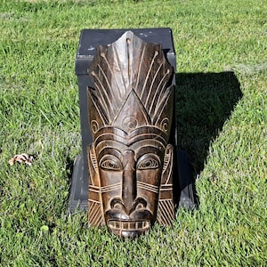 May include: A dark brown wooden wall mask featuring intricate hand-carved details, including a prominent headdress, almond-shaped eyes with circular pupils, a wide nose, and a mouth with visible teeth, all finished with a polished wood grain texture.