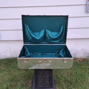 May include: An open, vintage green suitcase with a teal satin interior. The suitcase has brass-colored hardware and is sitting on a decorative black stand. The exterior shows signs of wear.