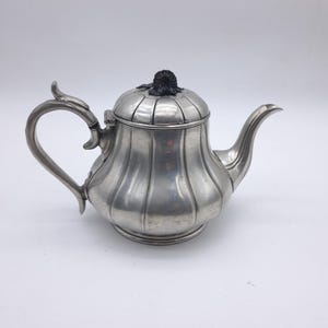 Puede incluir: Una tetera plateada con asa y boquilla curvadas. La tetera tiene un cuerpo acanalado y una tapa con un pomo oscuro y texturizado. El diseño es clásico y elegante, adecuado para servir té.