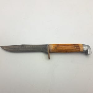 May include: A vintage hunting knife with a worn, silver blade and a handle featuring a light brown, textured design. The handle is accented with silver metal and two small rivets. The knife has a curved blade and a metal guard.