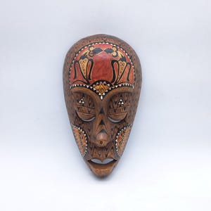 May include: A carved wooden wall mask featuring a natural wood grain finish with intricate black and white geometric patterns. The forehead area displays a smooth, reddish-brown section with stylised black figures, while the eyes and mouth are open.