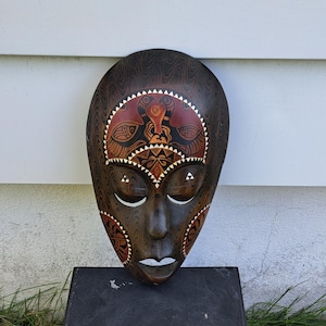 以下が含まれることがあります： 精巧な彫刻と塗装が施された、濃い茶色の木製マスク。マスクの中央には赤と茶色の模様があり、白いアクセントが施されています。顔は目を閉じ、穏やかな表情をしています。マスクは黒い面に展示されています。