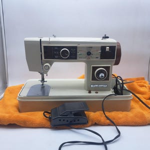 Peut inclure: Une machine à coudre Fleetwood vintage dans un boîtier couleur crème avec un panneau de commande noir présentant divers réglages de points et un cadran. La machine comprend une pédale noire avec un cordon d'alimentation et une plaque à aiguille.